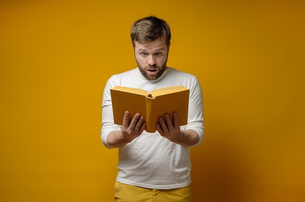 Attractive young man enthusiastically reads a book standing against a yellow background