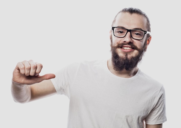 Attractive young man in eyeglasses pointing to his blank white tshirt with index finger showing empty space for your advertising text or logo statning isolated on white background