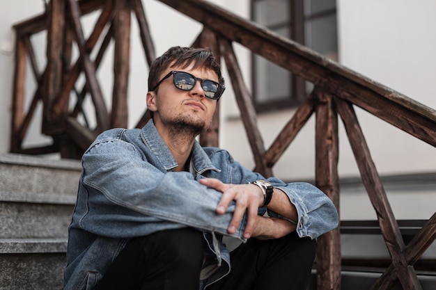 Photo attractive young man hipster in a fashionable blue denim jacket in stylish black sunglasses enjoys a weekend sitting on a vintage staircase in the city. trendy guy relaxes outdoors in a spring day.