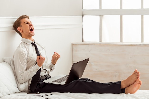 Attractive young man is using laptop while sitting on bed.