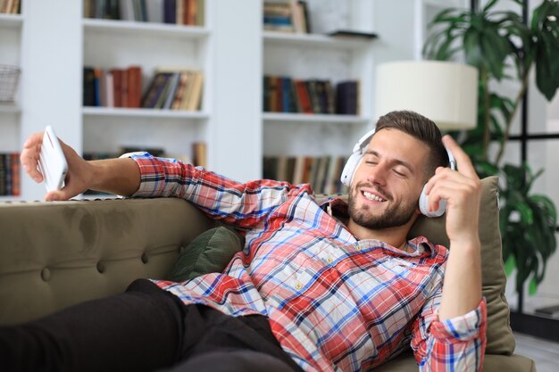 Attractive young man relaxing on a couch at home, listening music and playing on smartphone.