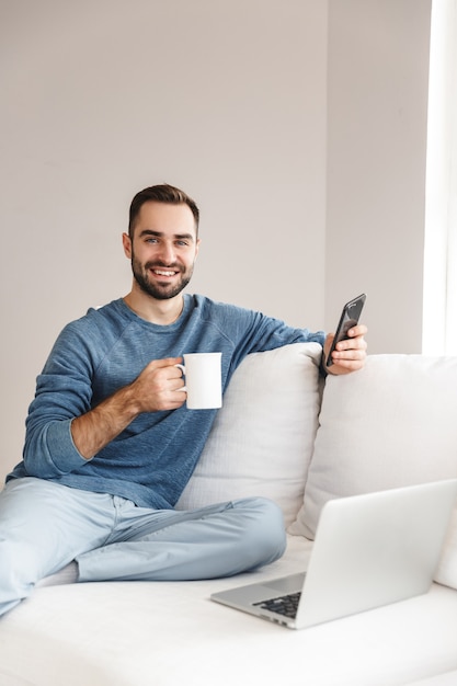 Attractive young man relaxing on a couch at home, using mobile phone, drinking coffee