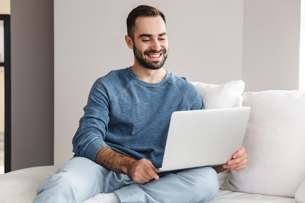 Attractive young man relaxing on a couch at home, working on laptop computer