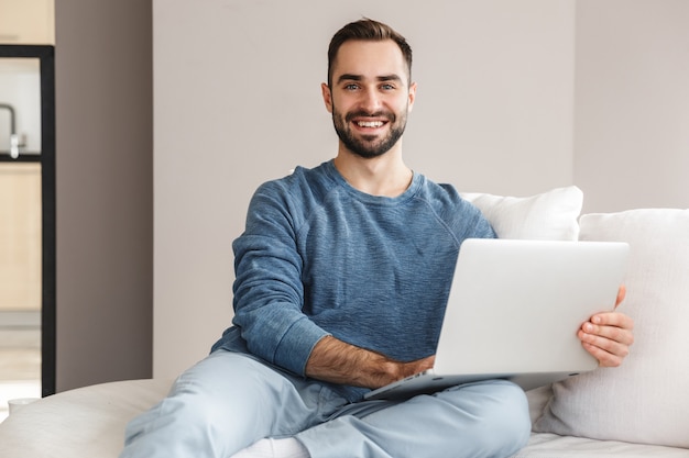 Giovane attraente che si rilassa su un divano a casa, lavorando su un computer portatile