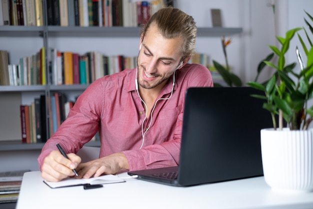 Фото Привлекательный молодой человек сидит за столом у себя дома, работая с ноутбуком, чувствуя себя счастливым
