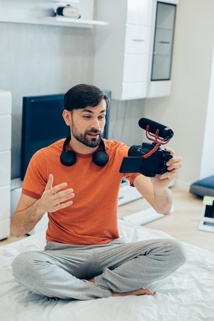 Photo attractive young man sitting crossed-legged on the bed with a modern camera and recording himself