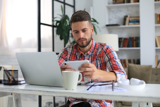 Attractive young man sitting at desk at home office and using mobile phone for cheking social nets.