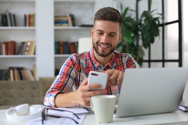 Attractive young man sitting at desk at home office and using mobile phone for cheking social nets.