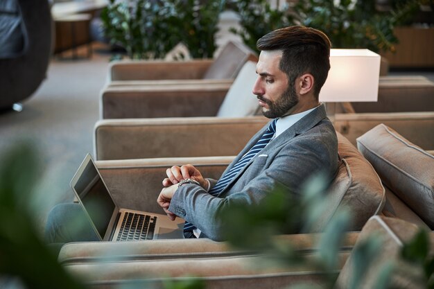 Attractive young man touching his watch while sitting in the armchair