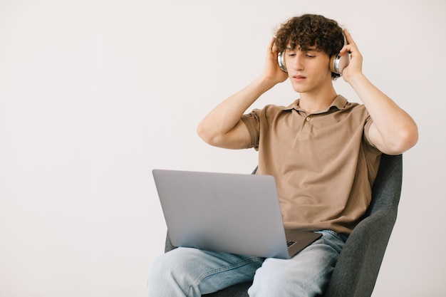 Attractive young man using laptop wearing headphones while sitting in armchair against white wall copy space Millennial male communicating online working or studying remotely on portable pc