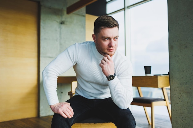 An attractive young man in a white sweater and trousers is smiling contentedly while sitting in a bright office Man in comfortable casual clothes