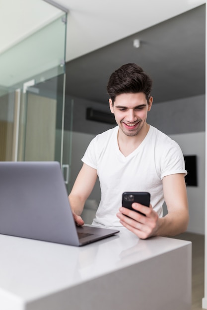 Привлекательный молодой человек, работающий из дома, сидя за столом на кухне, как он читает текстовое сообщение на своем мобильном телефоне