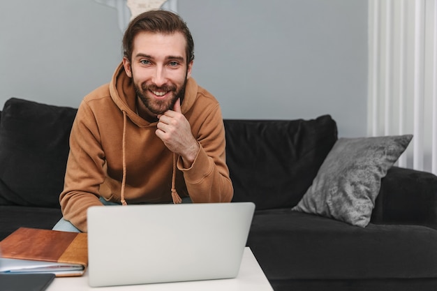 An attractive young man works at home using a laptop. Smiling guy with beard looking at the camera