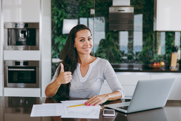 The attractive young modern business woman shows thumbs gesture cool and working with documents and laptop in the kitchen at home