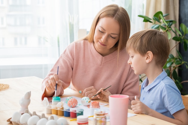 Attraente giovane madre e figlio che dipingono le uova con colori vivaci mentre si prepara per la pasqua