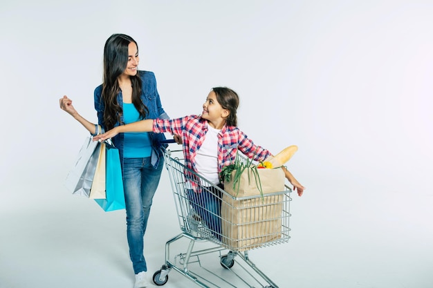 Attractive young mother with shopping bags is pushing her daughter who seat in food trolley with purchases and talking about something with smile