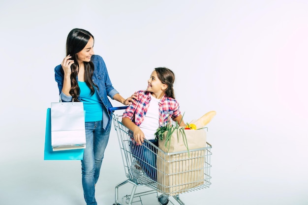 Attractive young mother with shopping bags is pushing her daughter who seat in food trolley with purchases and talking about something with smile