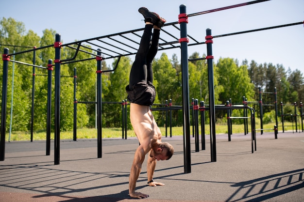 Attraente giovane atleta muscolare facendo verticale sul campo sportivo in giornata di sole con strutture