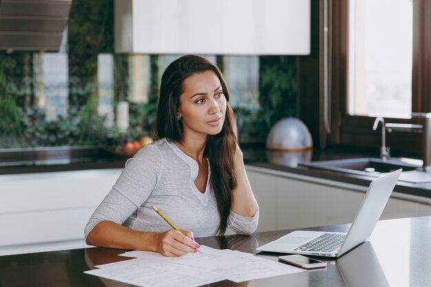 The attractive young pensive business woman working with documents and laptop in the kitchen at home