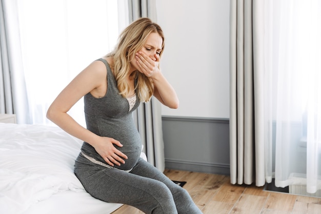 Attractive young pregnant blonde woman feeling sick while sitting on bed at home