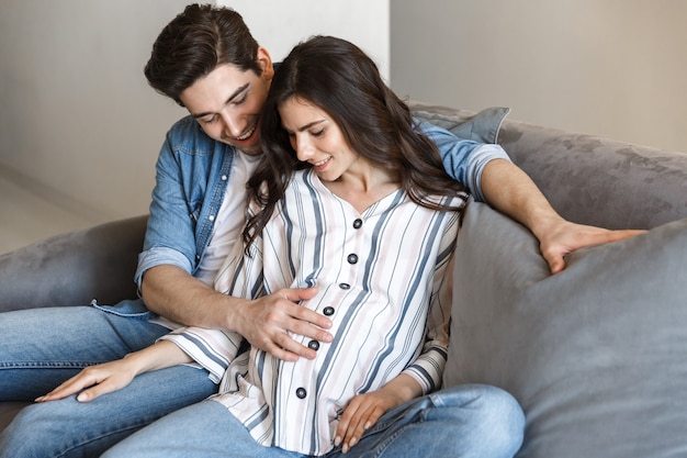 魅力的な若い妊娠中のカップルが自宅のソファでリラックスし、抱き締める