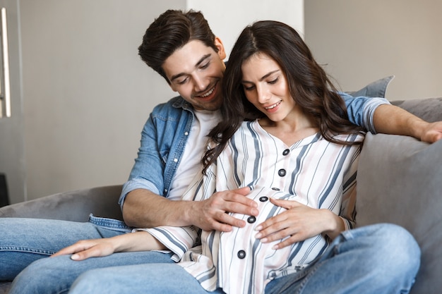 Attractive young pregnant couple relaxing on a couch at home, hugging