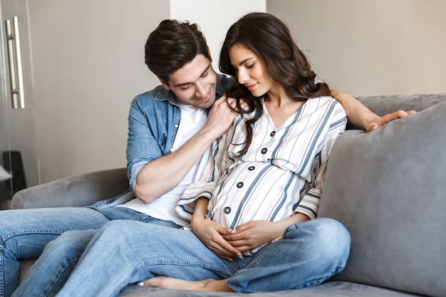 Attractive young pregnant couple relaxing on a couch at home, hugging