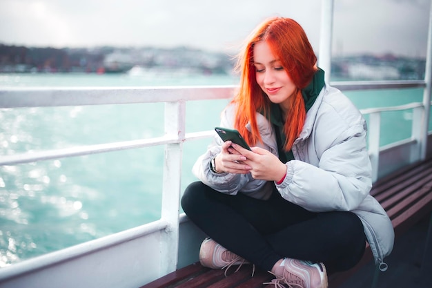 Photo attractive young redhead woman typing a message via her mobile phone in a ferryboat istanbul and she is smiling