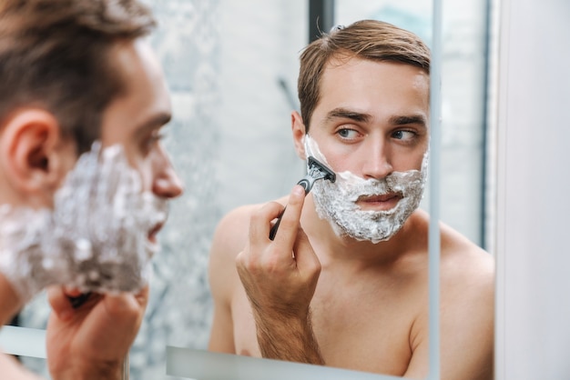 Attraente giovane uomo a torso nudo che si fa la barba mentre si trova allo  specchio del bagno