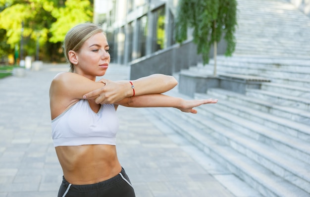 Привлекательная молодая стройная фитнес-женщина в спортивной одежде делает упражнения на растяжку и разминку для рук перед тренировкой на открытом воздухе