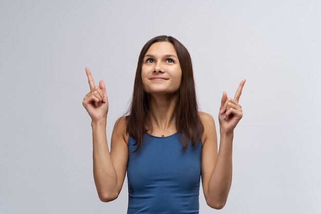 写真 タイトな青いtシャツと長いストレートの黒髪の魅力的な笑顔の若い女性の女の子は見上げると人差し指は上向き。広告スペースの概念と視線方向