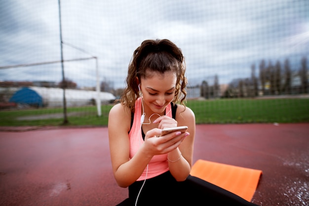 Привлекательная молодая sportive женщина проверяя телефон пока сидящ на оранжевой циновке.