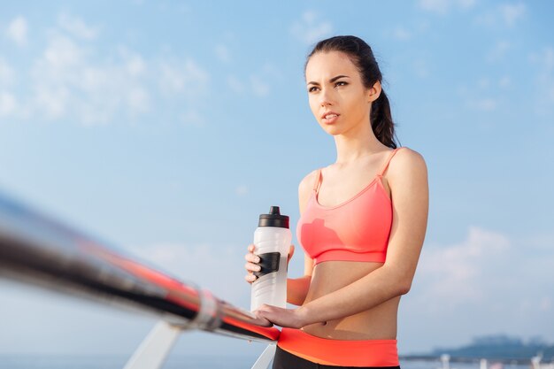 Attractive young sportswoman with bottle of water standing outdoors