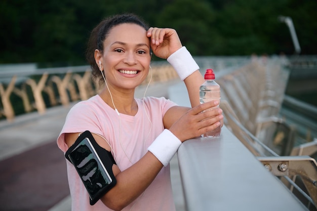 Attractive young sportswoman with smartphone holder on her arm, smiles with beautiful toothy smile looking at camera while resting on the city bridge after morning rung and physical exercises