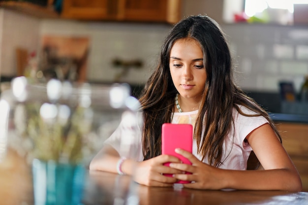 매력적인 10대 소녀가 실내에서 테이블에 앉아 쉬면서 휴대전화로 미디어를 읽거나 보고 있다