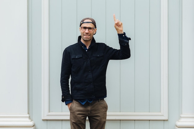 Attractive young unshaven male raises fore finger advertises something keeps hand in pocket wears stylish clothes poses alone against white wall has delighted expression Hey look there