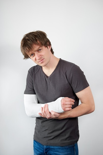 Attractive young upset caucasian man with plaster cast on his hand suffering from pain on a white wall