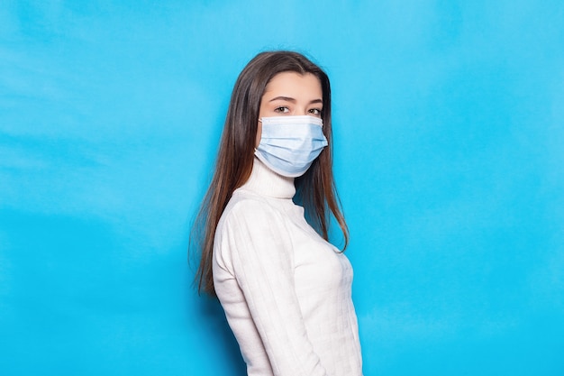 Attractive young woman 20s in face mask safe from coronavirus virus covid-19 during pandemic quarantine holding hands crossed isolated on bright blue colour background studio portrait