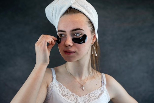 Attractive young woman applying collagen black eye patches under her eyes against a black wall