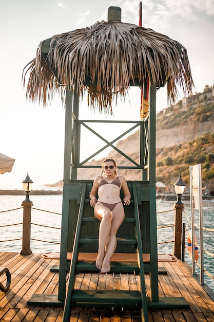 Attractive young woman in a beige bikini swimsuit at sunset sits in a wooden house by the sea