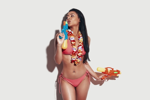 Attractive young woman in bikini blowing at a squirt gun while standing against grey background