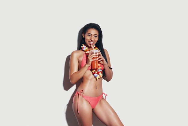 Привлекательная молодая женщина в бикини пьет освежающий коктейль и улыбается, стоя на сером фоне