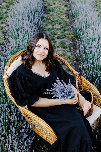 라벤더 밭으로 둘러싸인 의자에 앉아 꽃 바구니를 들고 검은 드레스를 입은 매력적인 젊은 여성