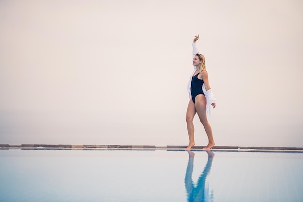 An attractive young woman in a black swimsuit and white shirt stands near the pool with a panoramic view