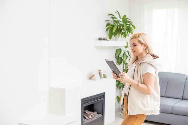 Attractive young woman in casual clothes is using a digital tablet, looking at camera and smiling at home.