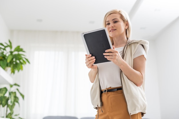 Attractive young woman in casual clothes is using a digital tablet, looking at camera and smiling at home.