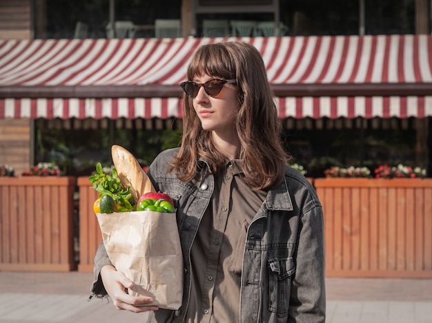 Attractive young woman in casual style clothes holding groceries bought from local vegetable and grocery store or market.