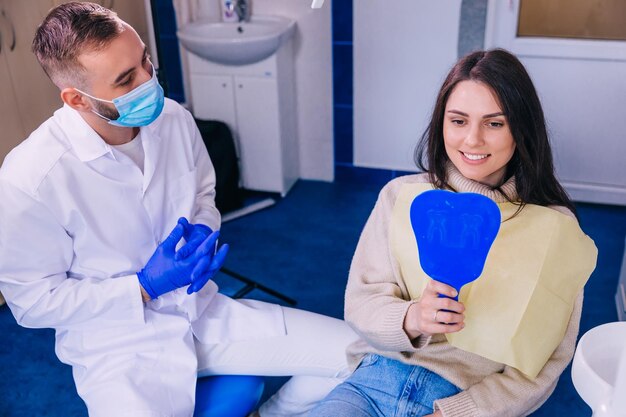 Attractive young woman checks her teeth in the mirror after dental treatment in the clinic