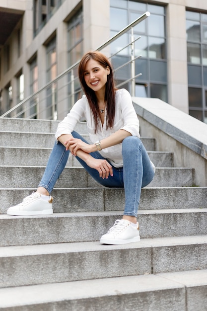 Foto attraente giovane donna in città. signora di affari che sta vicino al centro di affari. una donna d'affari stanca con una camicia bianca in blue jeans seduta si rilassa per strada vicino all'edificio di vetro del centro città