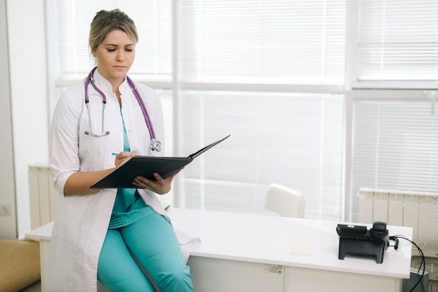 医者のクリップボードを保持している魅力的な若い女性医師。白衣と聴診器でクリップボードにメモを取りながら立っているHandome若い女性医師または看護師